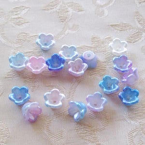 May include: A collection of small, flower-shaped beads in various pastel colors, including blue, pink, and purple. The beads have a pearlescent finish and are scattered on a light-colored fabric.