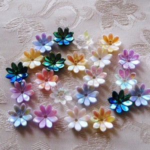 May include: Assortment of iridescent flower-shaped sequins in various colors, including pink, purple, blue, green, and yellow. The sequins are arranged in a circle on a white fabric background.