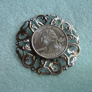 May include: A silver-toned metal pendant featuring a decorative filigree design with a United States quarter dollar coin in the center. The coin features the profile of George Washington and the words "United States of America, In God We Trust, Liberty, Quarter Dollar".