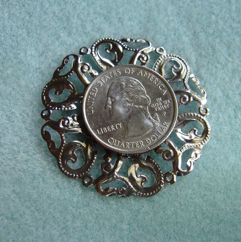 May include: A silver-toned metal pendant featuring a decorative filigree design with a United States quarter dollar coin in the center. The coin features the profile of George Washington and the words "United States of America, In God We Trust, Liberty, Quarter Dollar".