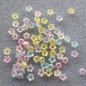 May include: A collection of small, flower-shaped beads in pastel colors, including pink, yellow, and blue. The beads have a hole in the center and are scattered on a light gray surface. Ideal for jewelry making and crafts.