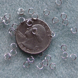 May include: A close-up of a silver US quarter with a pile of silver jump rings scattered around it. The jump rings are small and have a shiny finish.
