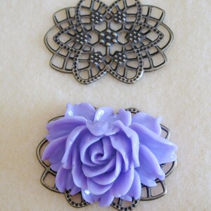 K&ouml;nnte beinhalten: Zwei dekorative Metallembellishments, eines ist ein filigranes Design und das andere ist ein Rahmen mit einer lavendelfarbenen Rose in der Mitte.