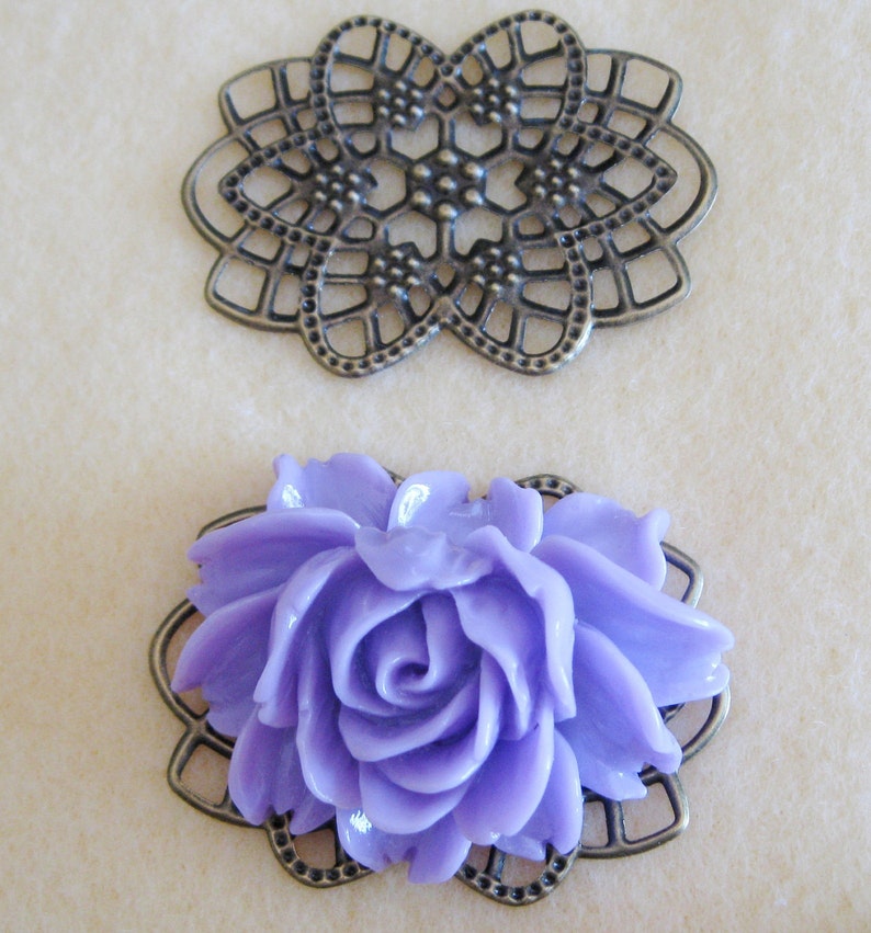K&ouml;nnte beinhalten: Zwei dekorative Metallembellishments, eines ist ein filigranes Design und das andere ist ein Rahmen mit einer lavendelfarbenen Rose in der Mitte.