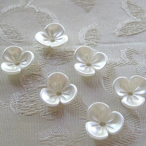 May include: Six white plastic flower-shaped buttons with a raised center and a hole in the middle. The buttons are arranged in a semi-circle on a white fabric background.