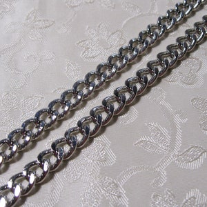 May include: Two silver chains with a curb link design. The chains are lying flat on a white fabric background.