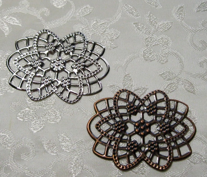 K&ouml;nnte beinhalten: Zwei dekorative filigranartige Metallst&uuml;cke, eines silberfarben und eines kupferfarben, mit einem floralen Design. Die St&uuml;cke sind filigran und detailliert, mit einem zarten Muster aus Kreisen und Linien.