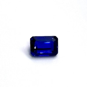 Puede incluir: Una gema rectangular de color azul oscuro con talla esmeralda. La gema se encuentra sobre un fondo blanco, mostrando su color vibrante y facetas. La forma de la piedra es un rectángulo clásico con esquinas cortadas.