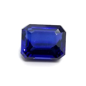 Puede incluir: Una gema azul oscuro tallada en forma rectangular. La gema tiene una superficie brillante y reflectante, sobre un fondo blanco. Las facetas crean un juego de luces y sombras, realzando su brillo.