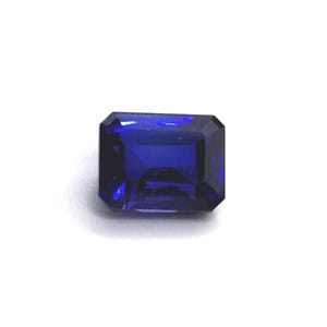 Puede incluir: Una gema rectangular de color azul oscuro con talla esmeralda. La gema tiene una superficie facetada que refleja la luz, creando un brillo brillante. La piedra se encuentra sobre un fondo blanco, resaltando su color y claridad.