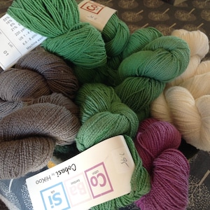 May include: A collection of yarn skeins in various colors, including green, gray, cream, and purple. The yarn is labeled with details about the fiber content and care instructions. The image shows a variety of yarn for knitting or crocheting projects.