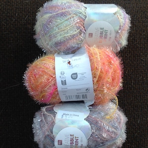 以下が含まれることがあります： ピンク、オレンジ、シルバーの色合いのクリエイティブバブルプリントの糸が3玉。糸は、メタリックなストランドを持つふわふわした質感です。各ボールには製品情報が記載されたラベルが付いています。