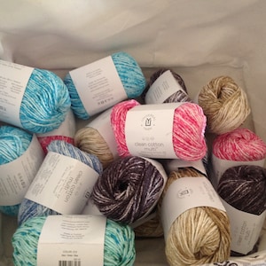 May include: A basket filled with various skeins of yarn in different colors and patterns, including blue, pink, brown, and beige. The yarn is labeled "Clean Cotton Multi" and is likely for knitting or crocheting.
