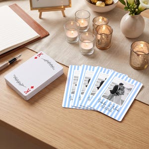 May include: A deck of playing cards with a King of Diamonds design and a stack of custom cards featuring light blue vertical stripes, a black and white photograph of a couple, and the text Isabella & Noah 11.23.2026 printed on the front.