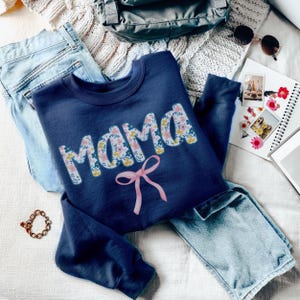 May include: A navy blue sweatshirt with the word "Mama" in a floral print design. A pink bow is below the word. The sweatshirt is laid on top of a pair of blue jeans.