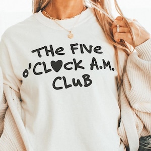 Puede incluir: Una sudadera blanca con el texto "The Five O'Clock A.M. Club" en letras negras con un corazón reemplazando la "O".