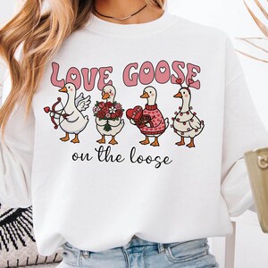 Puede incluir: Sudadera blanca con el texto "Love Goose on the Loose" en rosa. El gráfico presenta cuatro gansos de dibujos animados, cada uno con accesorios temáticos de San Valentín, como un arco y flechas, flores y luces.