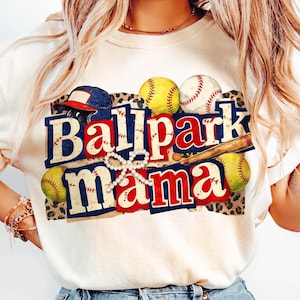 Può includere: T-shirt color crema con la scritta "Ballpark Mama" in lettere rosse, bianche e blu. Il design include palline da baseball, una mazza e un casco. La maglietta è abbinata a jeans azzurri.