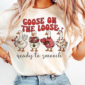 Puede incluir: Camiseta color crema con el texto rojo "GOOSE ON THE LOOSE" y cuatro gansos de dibujos animados. Cada ganso está decorado con motivos de San Valentín, con el texto "ready to smooch" debajo.