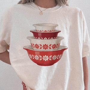 May include: Cream-colored t-shirt featuring a stack of four red and white floral patterned mixing bowls. The bowls are in varying sizes, with the largest at the bottom. The shirt is paired with red and white checkered shorts.