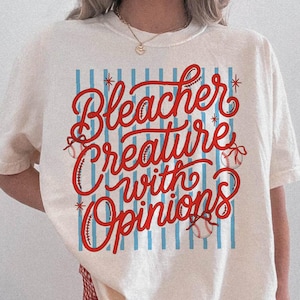 May include: Cream-colored t-shirt with the text "Bleacher Creature with Opinions" in red script. The design includes baseballs and blue vertical stripes. The shirt is paired with red and white checkered shorts.