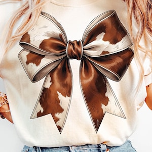 May include: A white t-shirt featuring a brown and white cow print bow on the front.