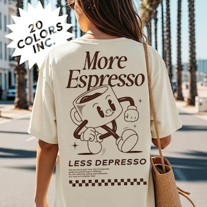 Könnte beinhalten: Cremefarbenes T-Shirt mit den braunen Aufschriften "More Espresso" und "Less Depresso". Das Shirt zeigt eine Cartoon-Kaffeetasse. Der Text "20 Colors Inc." ist ebenfalls sichtbar.