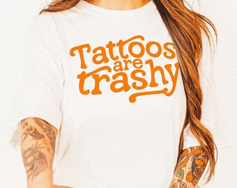 May include: White long-sleeve t-shirt with the orange text "Tattoos are trashy." The shirt is worn with a pair of black and brown cowboy boots with a star design. The person has multiple tattoos on their arms and legs.