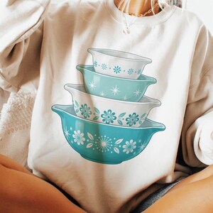 May include: A cream-colored sweatshirt featuring a stack of turquoise and white floral patterned bowls. The person is holding a black mug. The sweatshirt is a casual, comfortable style.