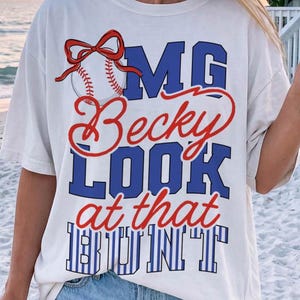 Può includere: T-shirt bianco con grafica di baseball e la scritta "OMG Becky LOOK at that BUNT" in blu e rosso. La grafica del baseball include un fiocco rosso e dettagli di cucitura.