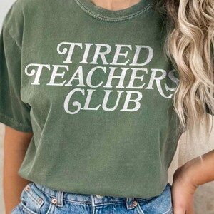 Könnte beinhalten: Ein grünes T-Shirt mit dem Text "TIRED TEACHERS CLUB" in weißen Buchstaben.