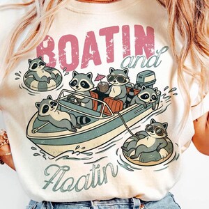 Może przedstawiać: Kremowa koszulka z kreskówkową ilustracją szopów pływających łódką i unoszących się na wodzie. Projekt zawiera łódź z szopami, niektóre w okularach przeciwsłonecznych, a inne w kołach ratunkowych. Tekst brzmi "BOATIN and Floatin".