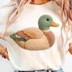 May include: Cream-colored t-shirt featuring a knitted duck design in shades of green, brown, and tan. The duck is the focal point, with detailed stitching. The shirt is paired with light blue denim jeans.