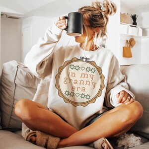 Puede incluir: Sudadera color crema con un dise&ntilde;o de punto de cruz que dice "in my granny era" en un marco circular. El dise&ntilde;o incluye motivos de coraz&oacute;n y estrella verdes y rosas. Una taza negra se sostiene frente a la cara.