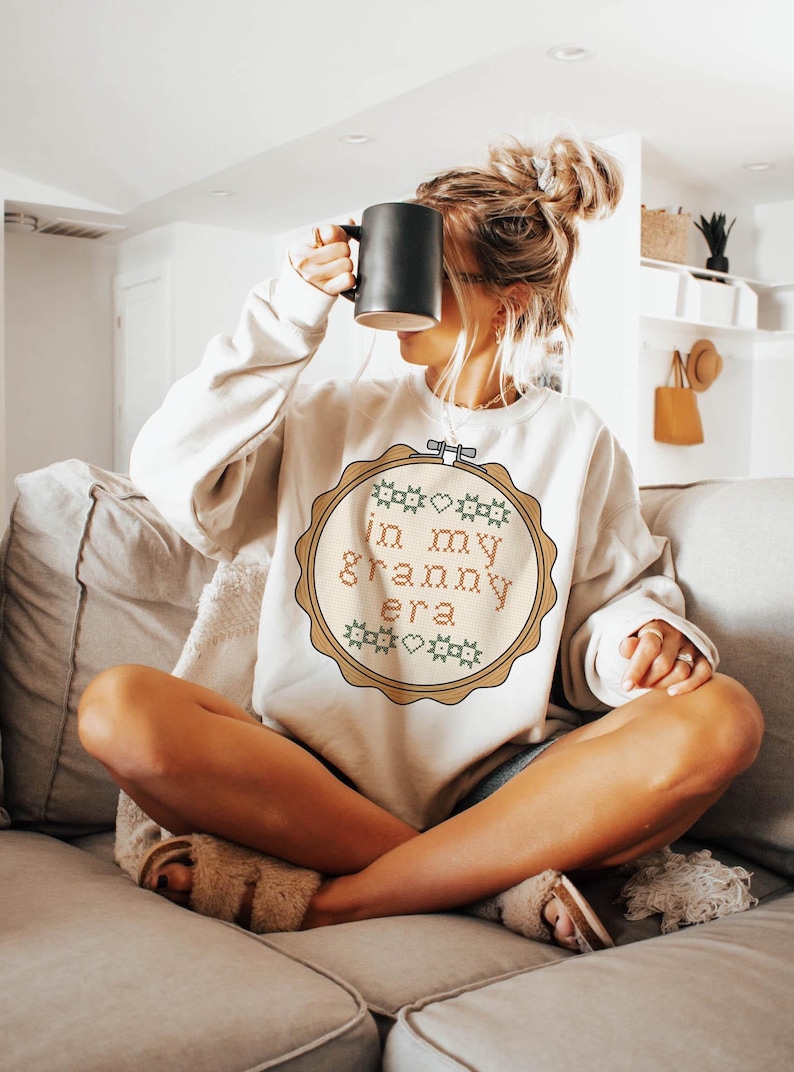 Puede incluir: Sudadera color crema con un dise&ntilde;o de punto de cruz que dice "in my granny era" en un marco circular. El dise&ntilde;o incluye motivos de coraz&oacute;n y estrella verdes y rosas. Una taza negra se sostiene frente a la cara.