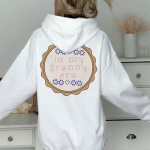 Puede incluir: Sudadera con capucha blanca con un dise&ntilde;o bordado circular en la espalda. El dise&ntilde;o presenta el texto "in my granny era" en punto de cruz rosa y azul, rodeado por un borde marr&oacute;n.