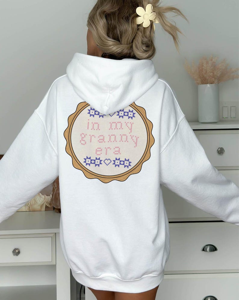 Puede incluir: Sudadera con capucha blanca con un dise&ntilde;o bordado circular en la espalda. El dise&ntilde;o presenta el texto "in my granny era" en punto de cruz rosa y azul, rodeado por un borde marr&oacute;n.