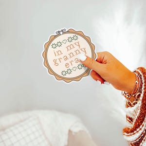 Puede incluir: Una pegatina redonda con un dise&ntilde;o de punto de cruz. La pegatina presenta la frase "in my granny era" en marr&oacute;n claro, rodeada de elementos decorativos verdes y marrones. La pegatina est&aacute; sujeta por una mano con esmalte de u&ntilde;as blanco.