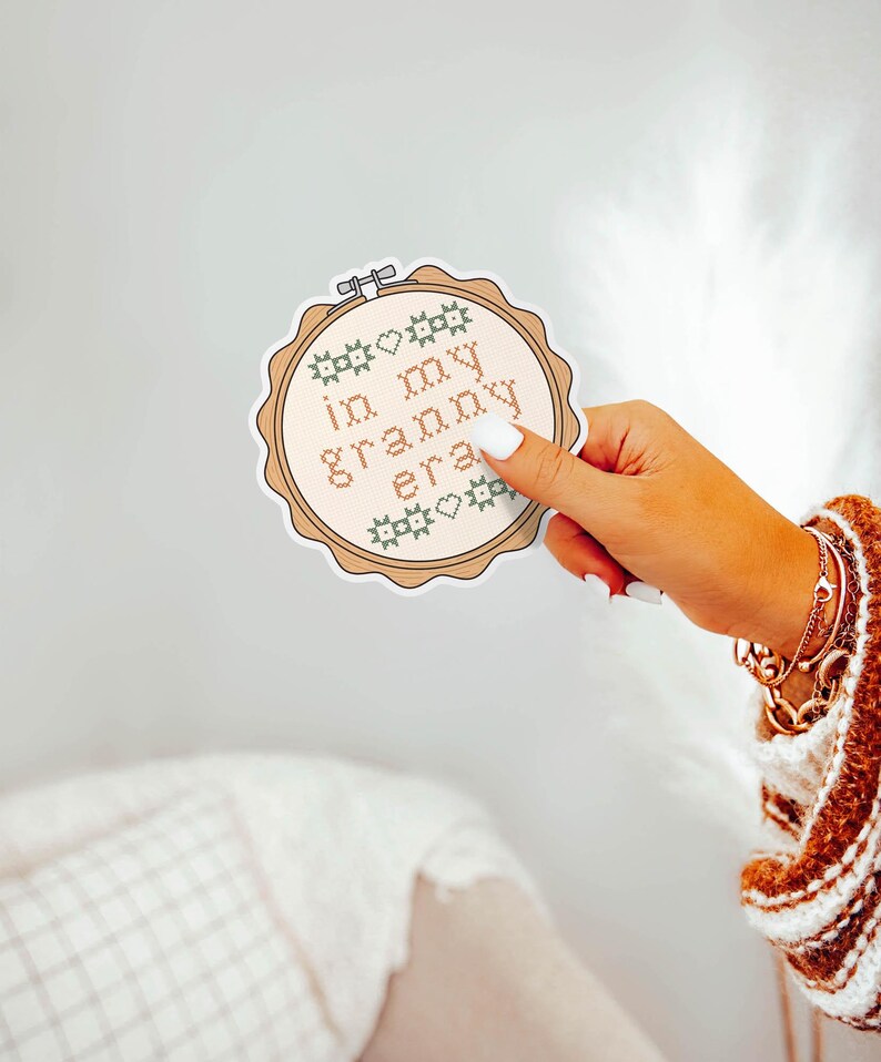 Puede incluir: Una pegatina redonda con un dise&ntilde;o de punto de cruz. La pegatina presenta la frase "in my granny era" en marr&oacute;n claro, rodeada de elementos decorativos verdes y marrones. La pegatina est&aacute; sujeta por una mano con esmalte de u&ntilde;as blanco.