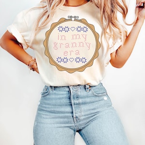 Puede incluir: Camiseta color crema con un dise&ntilde;o de punto de cruz dentro de un bastidor de bordado. El dise&ntilde;o incluye el texto "in my granny era" en letras rosas y azules. La camiseta se combina con vaqueros azul claro.