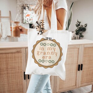 Puede incluir: Una bolsa de mano de lona blanca con un dise&ntilde;o de punto de cruz que dice "in my granny era" en marr&oacute;n y verde. La bolsa tiene un dise&ntilde;o de aro de bordado de madera marr&oacute;n alrededor del texto. La bolsa tiene una correa de color crema.