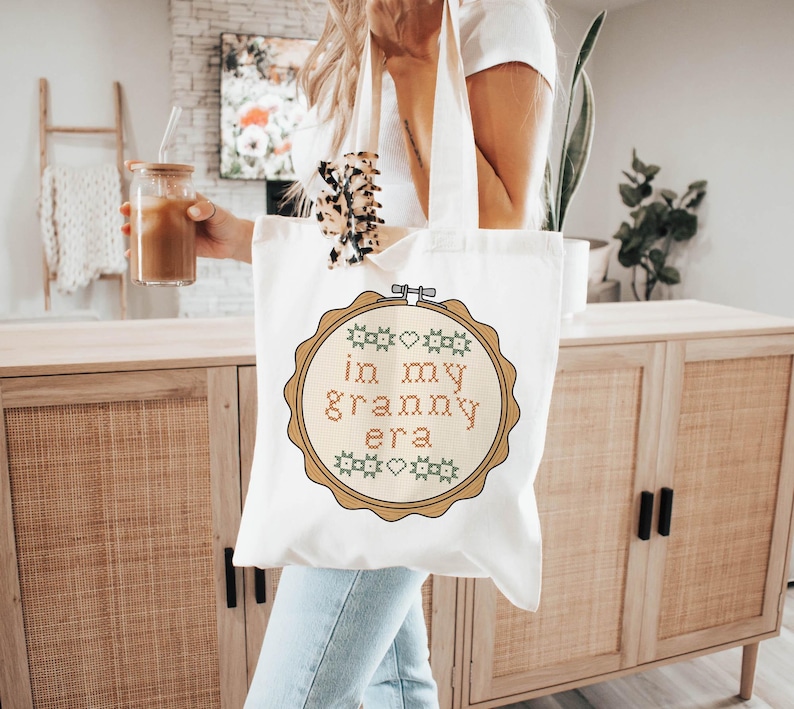Puede incluir: Una bolsa de mano de lona blanca con un dise&ntilde;o de punto de cruz que dice "in my granny era" en marr&oacute;n y verde. La bolsa tiene un dise&ntilde;o de aro de bordado de madera marr&oacute;n alrededor del texto. La bolsa tiene una correa de color crema.