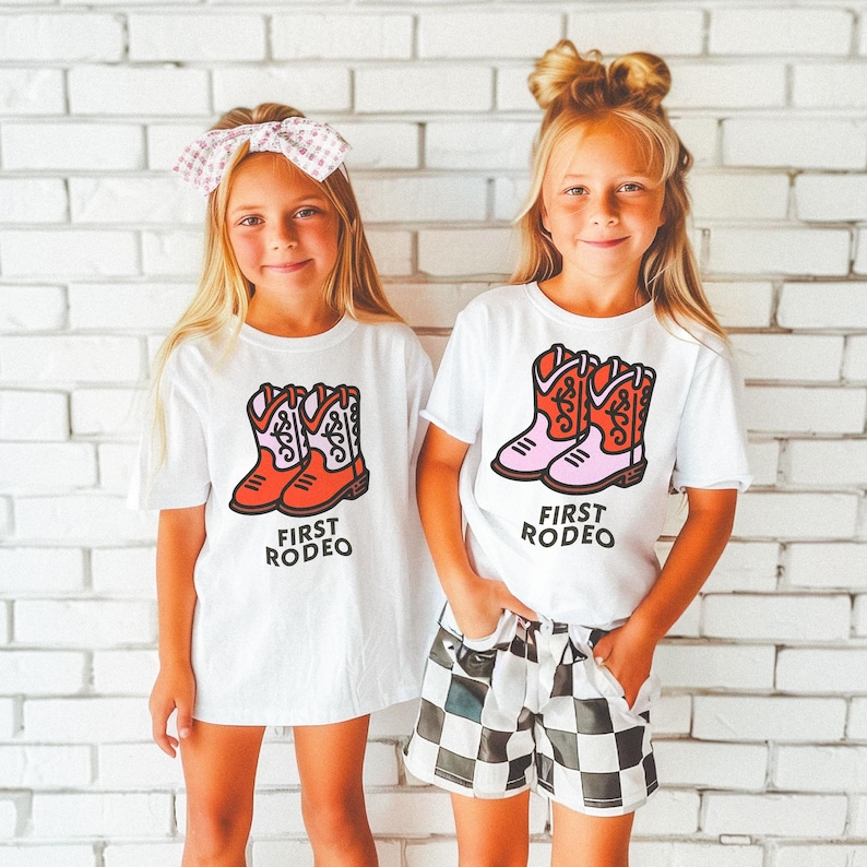May include: Two children wearing white t-shirts with a graphic of red cowboy boots and the text "First Rodeo". The girl on the left is wearing a pink bow in her hair. The girl on the right is wearing black and white checkered shorts.