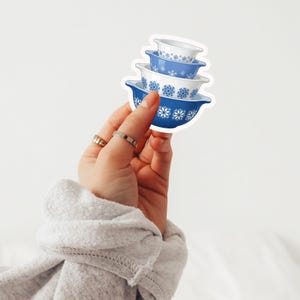 May include: A sticker featuring a stack of four blue and white mixing bowls with snowflake designs. The bowls are in varying sizes, with the largest at the bottom and the smallest at the top. The sticker is held by a hand.