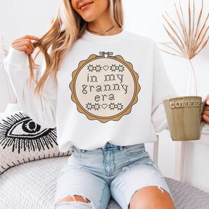 Puede incluir: Sudadera blanca con un dise&ntilde;o de punto de cruz en un bastidor de bordado de madera. El dise&ntilde;o dice "in my granny era" en una fuente de punto de cruz. La sudadera se combina con vaqueros azul claro. Se sostiene una taza verde con la palabra "coffee".
