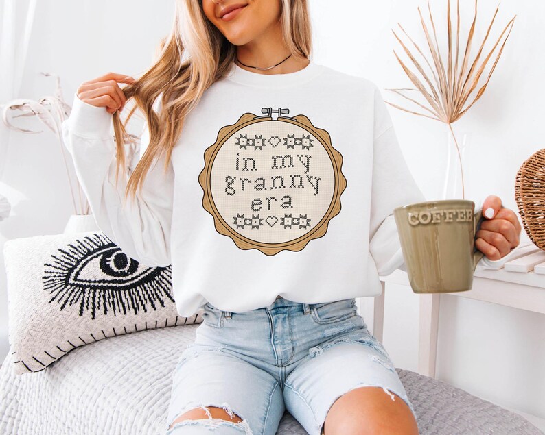 Puede incluir: Sudadera blanca con un dise&ntilde;o de punto de cruz en un bastidor de bordado de madera. El dise&ntilde;o dice "in my granny era" en una fuente de punto de cruz. La sudadera se combina con vaqueros azul claro. Se sostiene una taza verde con la palabra "coffee".