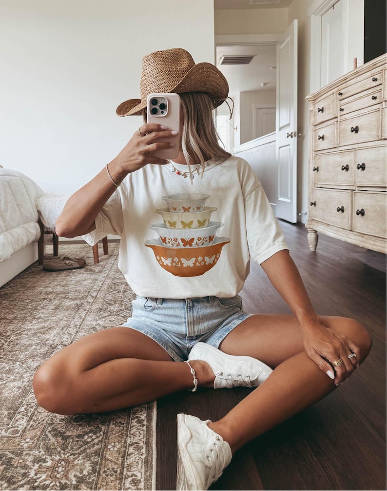May include: A white t-shirt featuring a stack of vintage-style mixing bowls with butterfly designs in orange, yellow, and white. The person is wearing a straw cowboy hat, denim shorts, and white sneakers. The person is sitting on a dark wood floor.