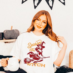 May include: White t-shirt with a vintage illustration of a witch flying on a broomstick. The text "Season of the Witch" is printed in orange and red.