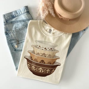 May include: Cream-colored t-shirt featuring a stack of vintage-style mixing bowls with a mushroom design. The shirt is paired with light-wash denim jeans, a gold necklace, and a tan hat. Dried flowers add a touch of color.