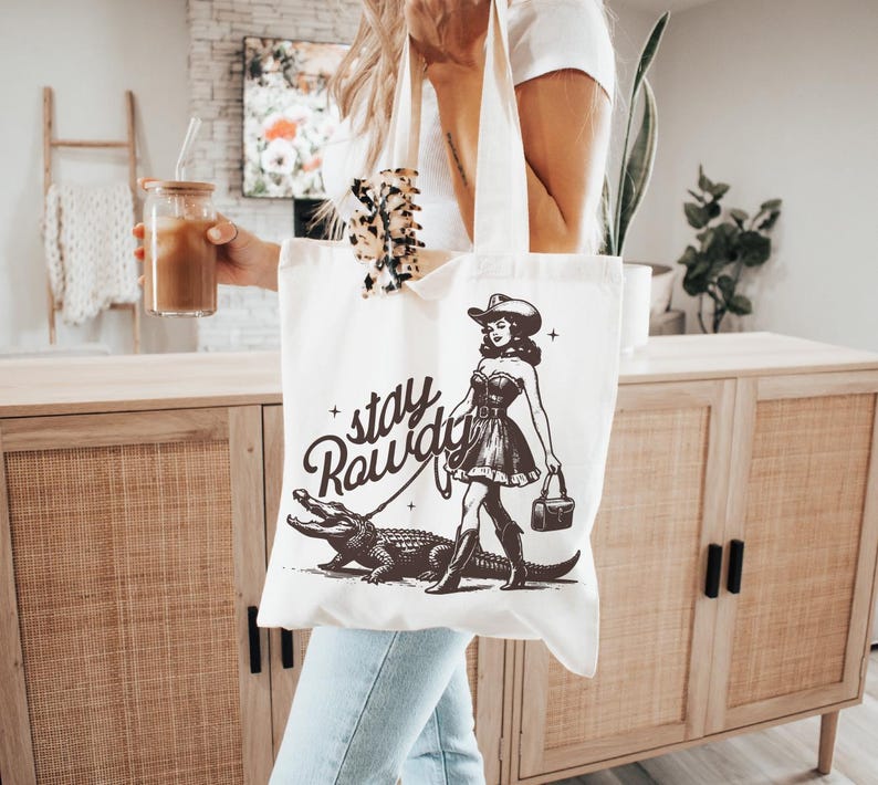 K&ouml;nnte beinhalten: Wei&szlig;e Canvas-Tragetasche mit einer Schwarzwei&szlig;-Illustration einer Frau im Cowboyhut, die ein Krokodil spazieren f&uuml;hrt. Der Text "stay rowdy" ist auf der Tasche aufgedruckt.